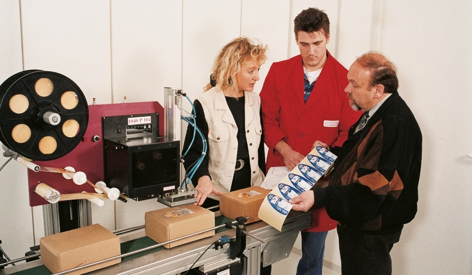 Zwei Männer und eine Frau an einer Etikettieranlage. Sinnbild für die Herstellung von maßgeschneiderten Etikettier- und Sondermaschinen.